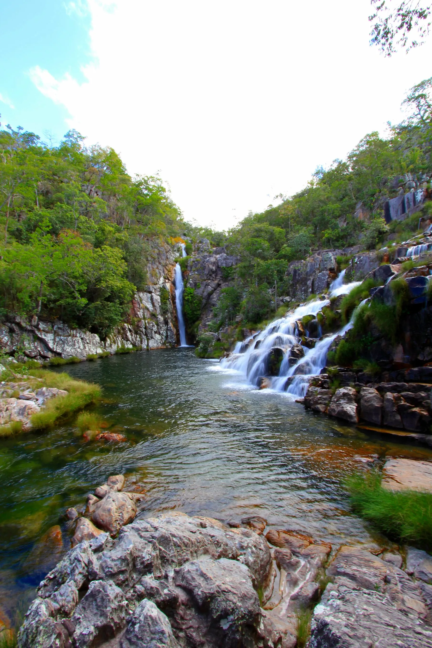 Cachoeira larga caindo em vários filetes sobre rocha escura, formando um poço verde cercado de mata