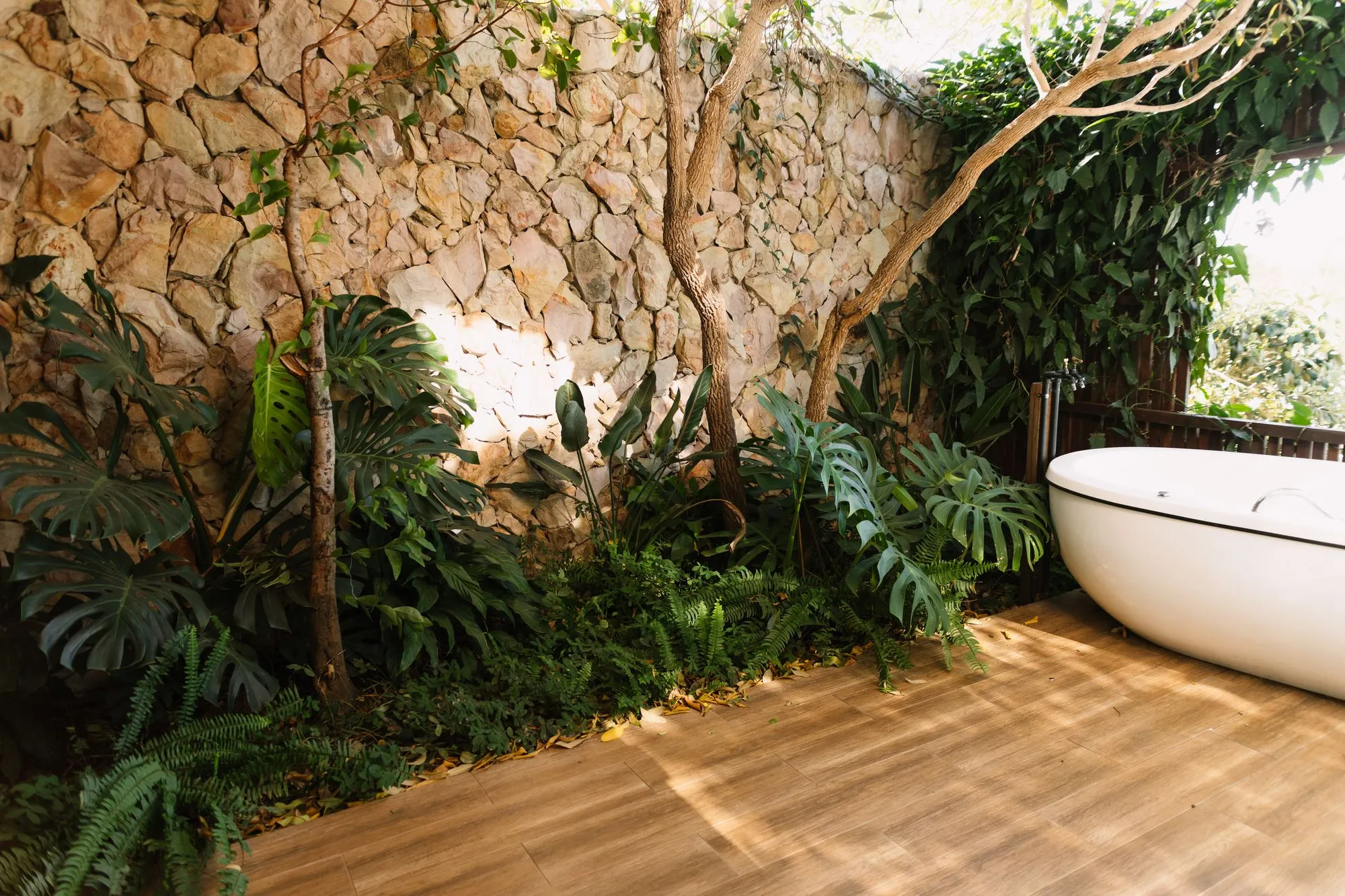 Banheira no deck de madeira do jardim privativo, entre um muro de pedra e plantas tropicais