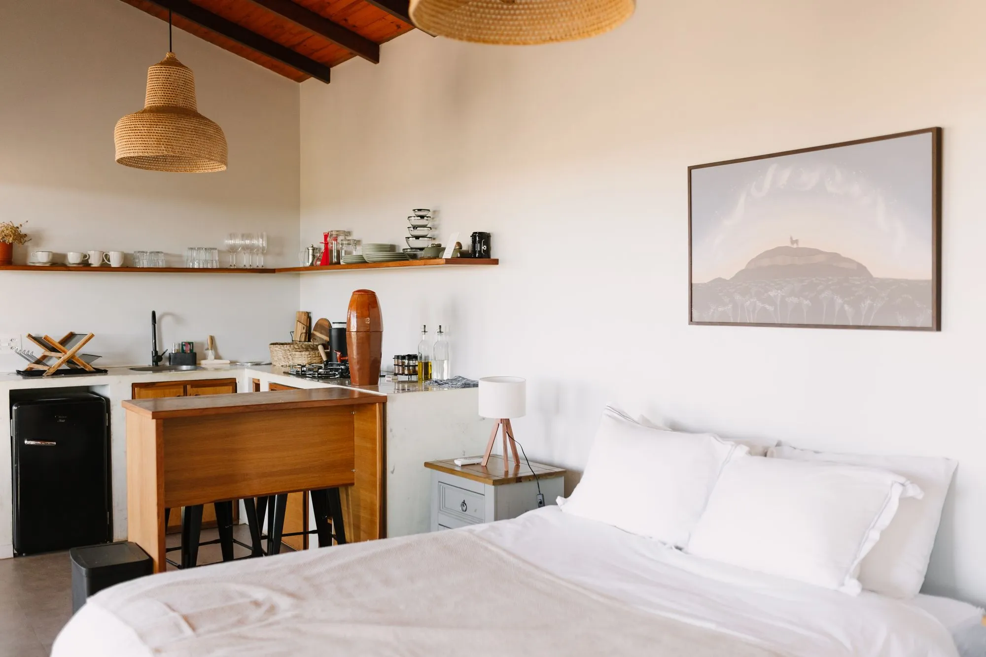 Cama queen com roupa de cama branca e, ao fundo, a cozinha com frigobar retrô e bancada de madeira