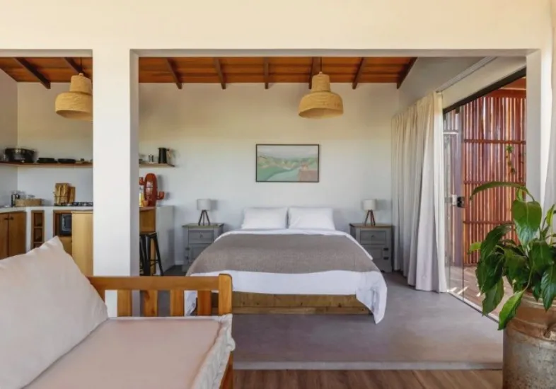 A cama vista da sala, com o sofá em primeiro plano e a porta de madeira ripada aberta