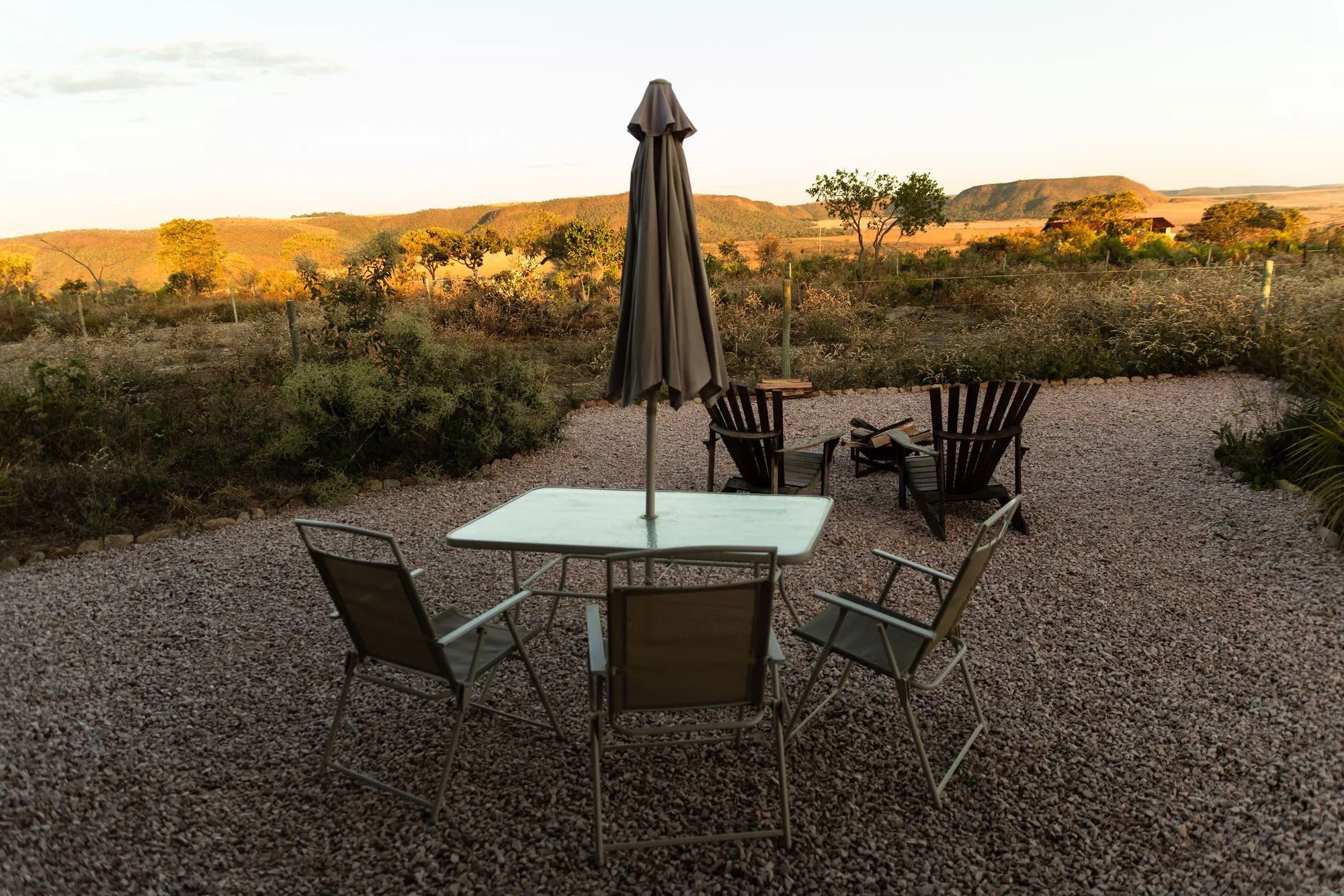 Área externa ao pôr do sol: mesa com guarda-sol, cadeiras e a fogueira diante das chapadas