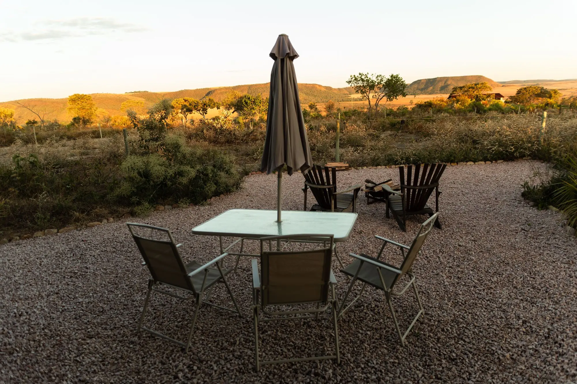 Área externa ao pôr do sol: mesa com guarda-sol, cadeiras e a fogueira diante das chapadas