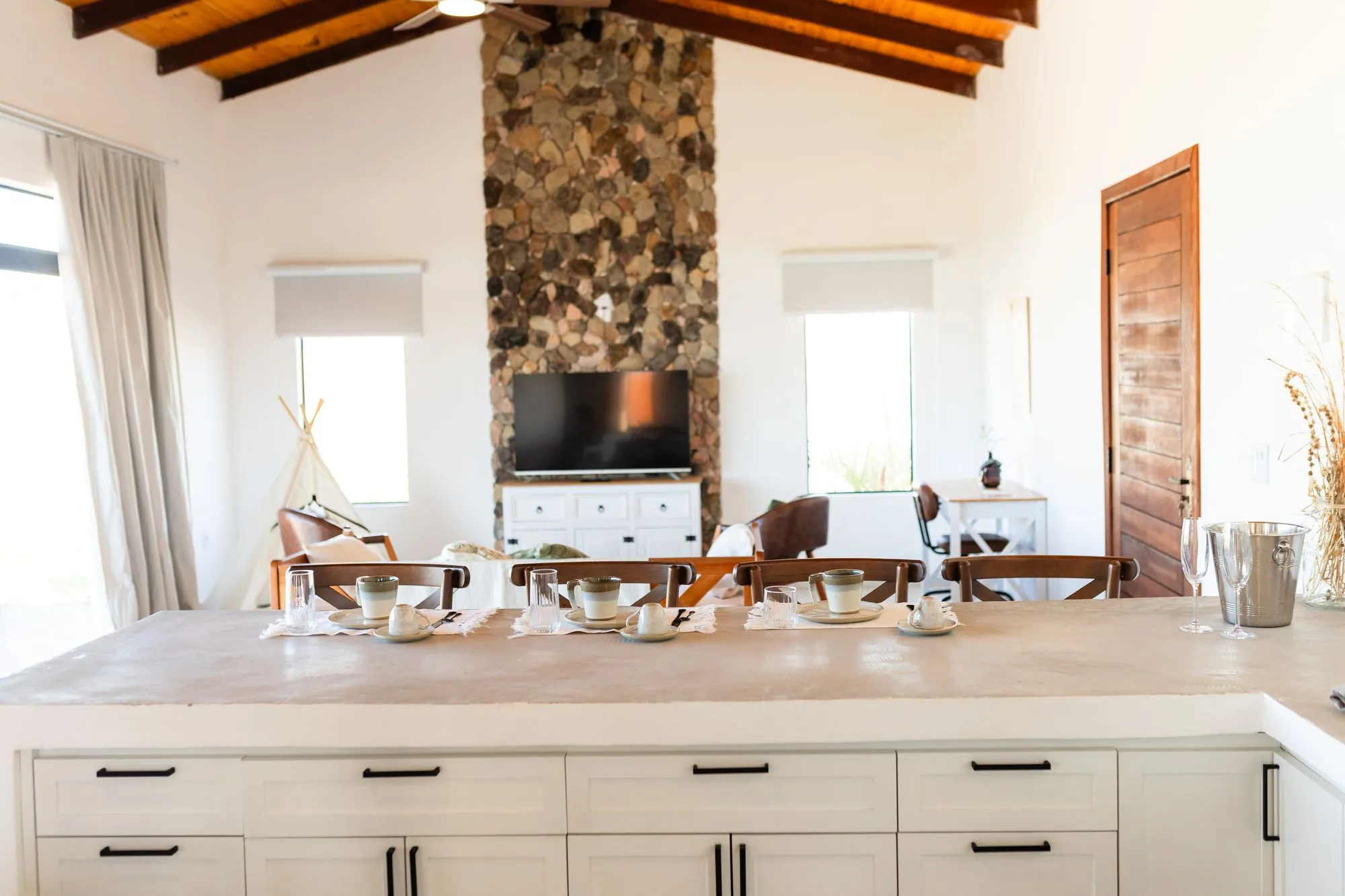 Sala com parede de pedra, TV e teto de madeira, vista por cima da bancada da cozinha