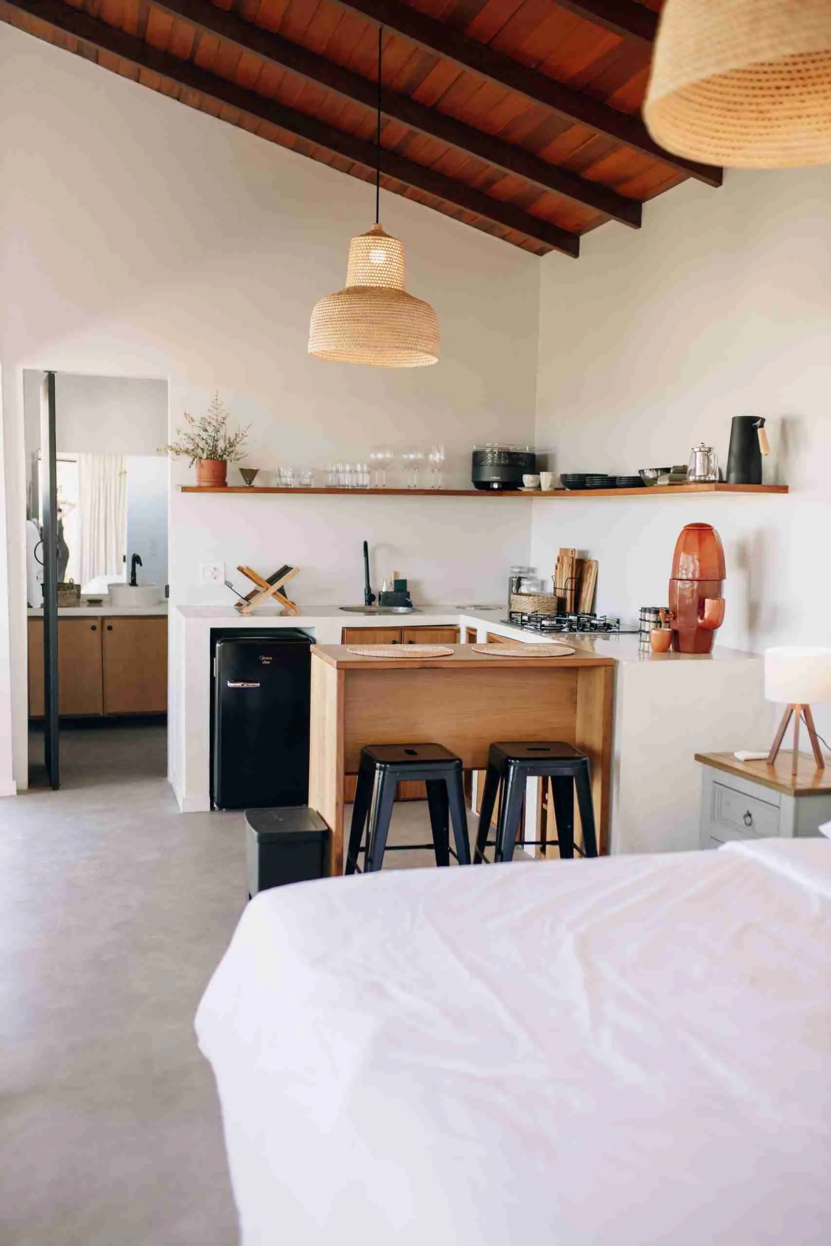 Full chalet interior with bed, wood-island kitchen, black stools, retro fridge and rattan pendant lights