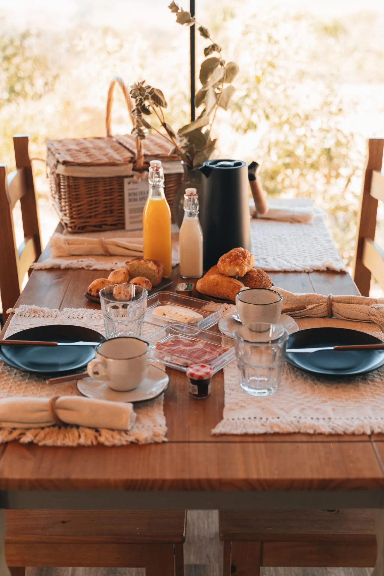 Mesa de café da manhã no chalé com cesta de vime, pães artesanais, suco de laranja, geleia e louça rústica
