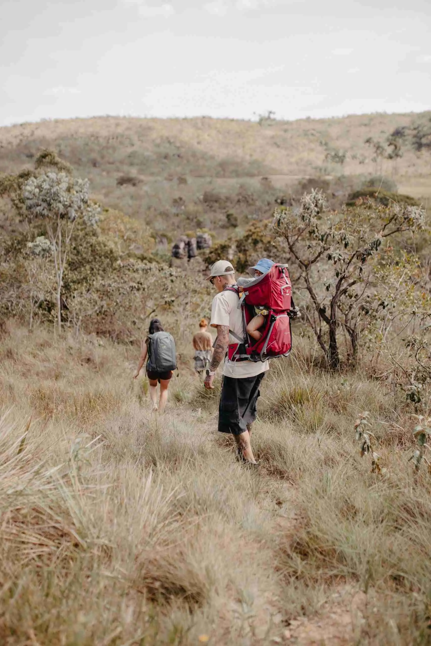 Família fazendo trilha pelo Cerrado, pai carregando bebê na mochila canguru vermelha