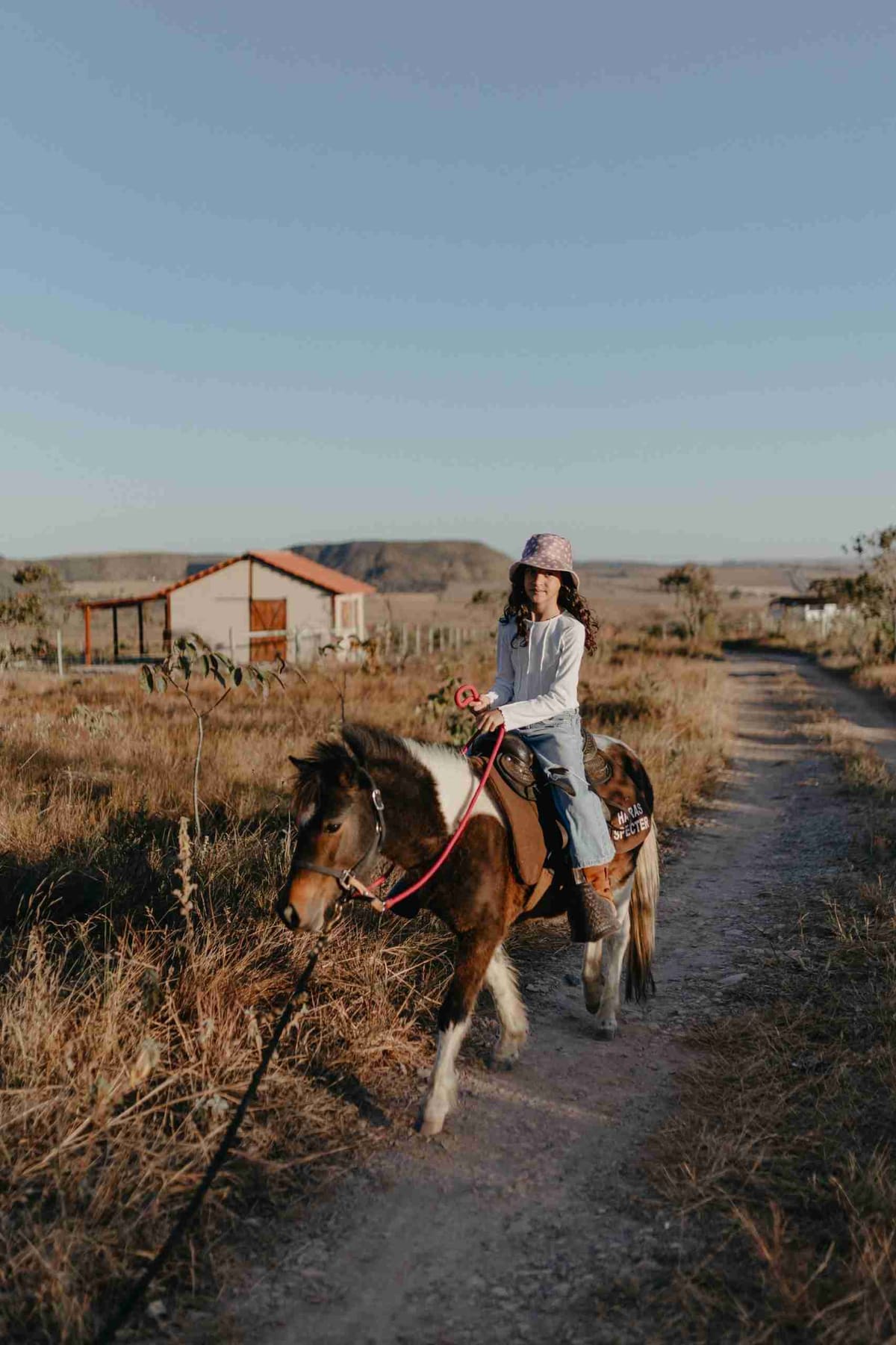 Criança sorridente montada em pônei malhado na trilha do rancho, com a chapada e um chalé ao fundo